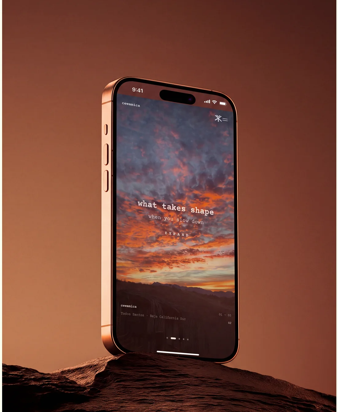 Un iPhone apoyado en una roca mostrando el sitio de Cewámica con un cielo de atardecer